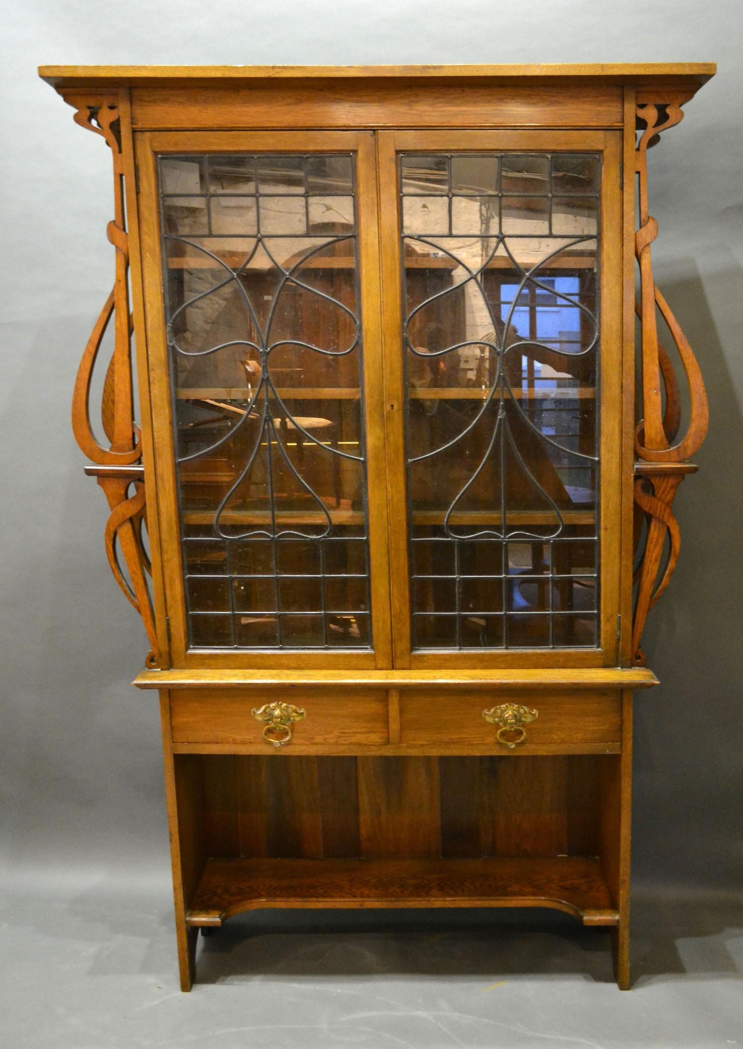 An Art Nouveau Oak Bookcase with two lead glazed doors enclosing shelves flanked by shaped pierced