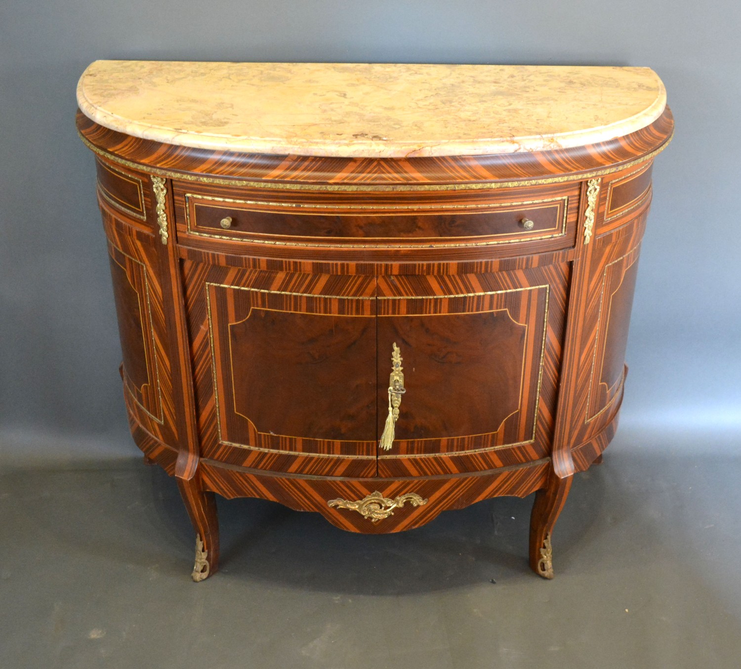 A French style demi-lune side cabinet, the variegated marble top above a frieze drawer with brass