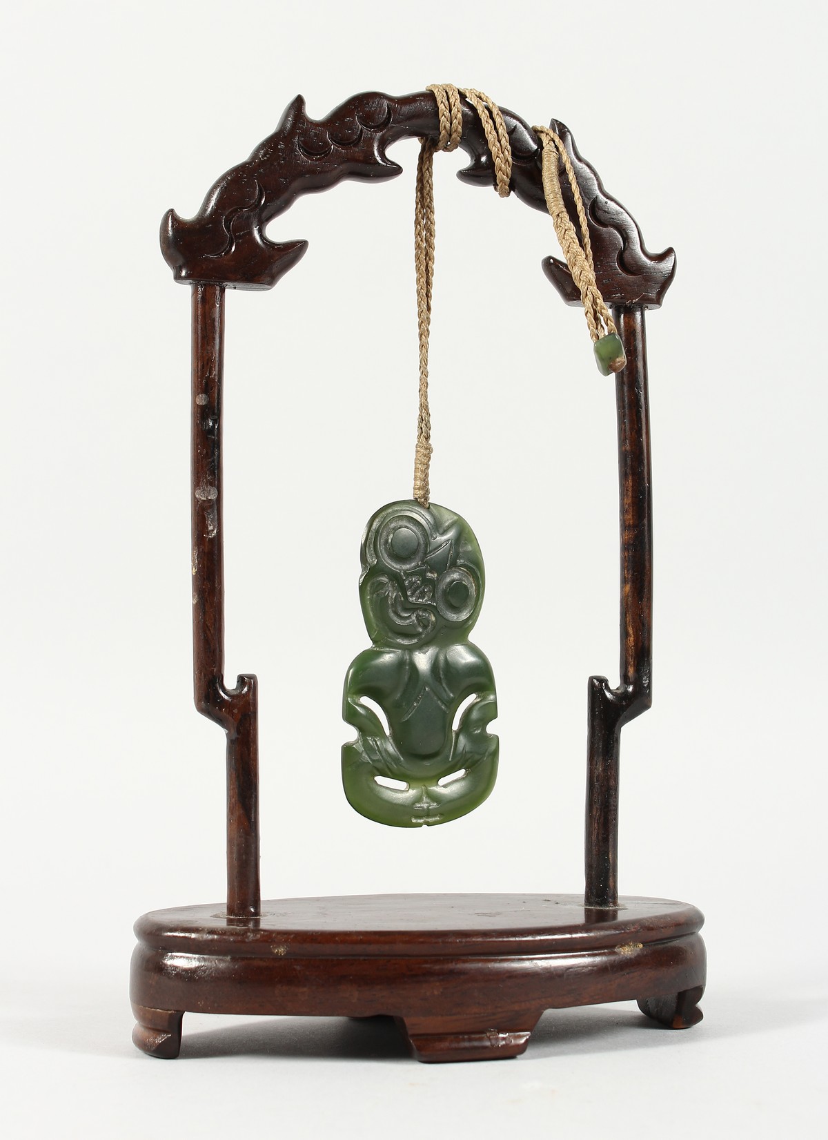 A NEW ZEALAND CARVED JADE TIKKI, suspended from a hardwood frame. Tikki: 2.75ins high.