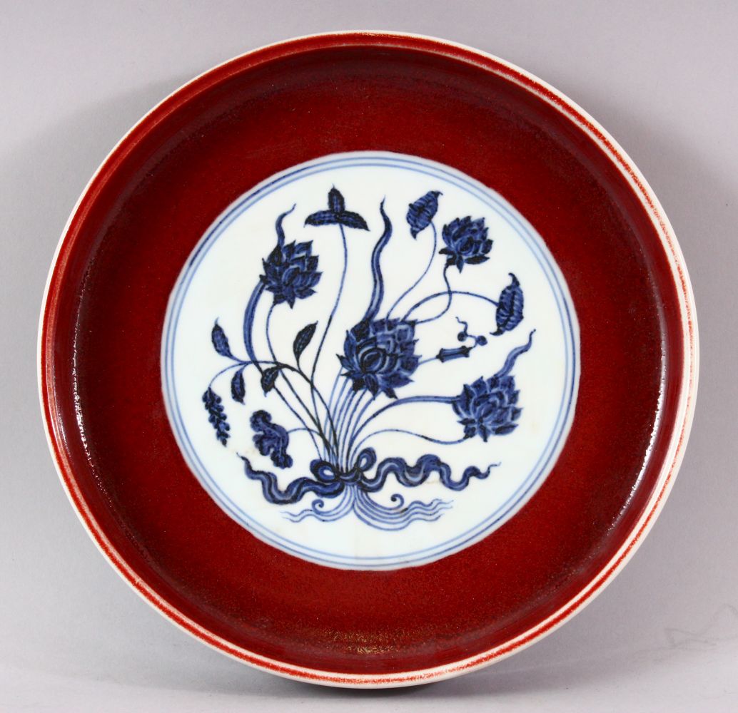 A MING STYLE COPPER RED CIRCULAR PORCELAIN DISH, the central blue and white panel painted with lotus