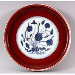 A MING STYLE COPPER RED CIRCULAR PORCELAIN DISH, the central blue and white panel painted with lotus
