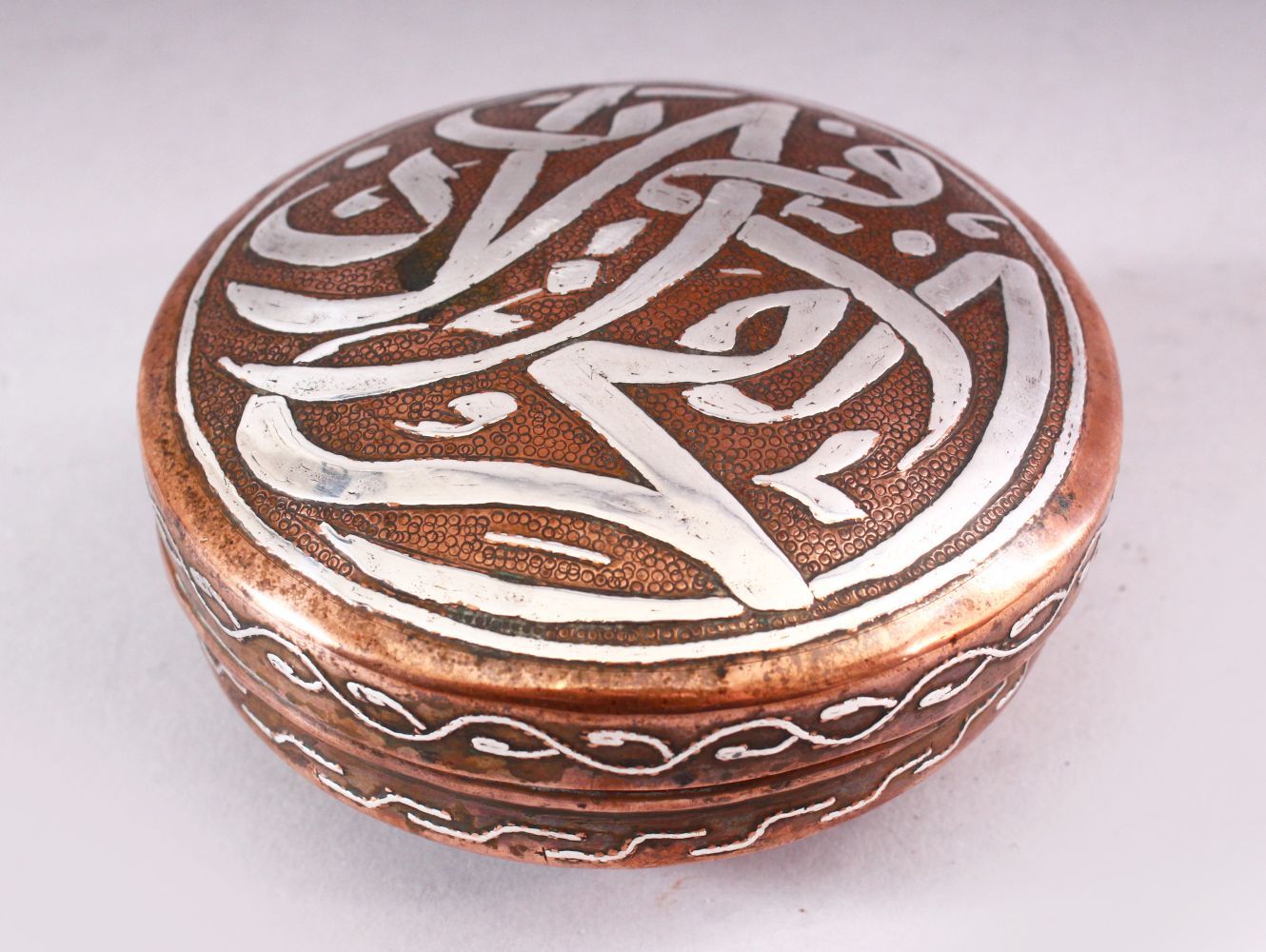 AN ISLAMIC CIRCULAR COPPER BOX AND COVER, the cover with silver inlaid calligraphy, 10cm diameter.