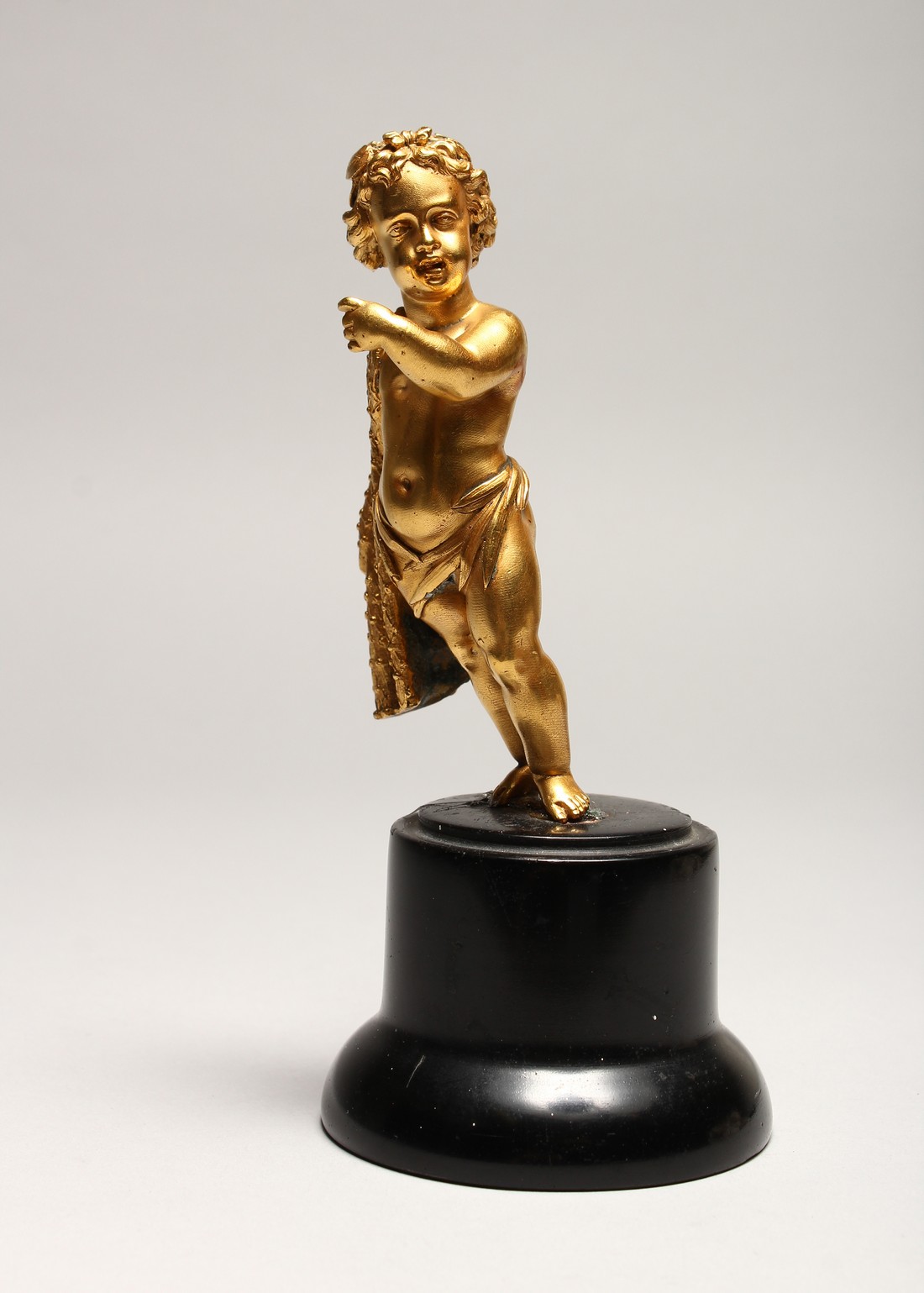 A GOOD SMALL GILDED METAL MODEL OF A CHERUB, standing with a cloak draped over one shoulder, on a