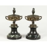 A PAIR OF BRONZE TWO HANDLED URNS AND COVERS on circular marble bases. 10ins high.