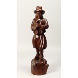 A CARVED WOOD FIGURE OF A MAN PLAYING A BLADDER PIPE. 19ins high.