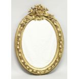 A DECORATIVE GILT FRAMED OVAL WALL MIRROR, the cresting with musical instruments, the frame with