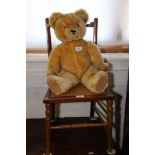 A gold plush teddy bear with growler, seated on an Edwardian caned seat chair