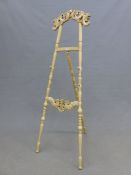 A CREAM PAINTED FLOOR STANDING EASEL, THE FOLIATE BRACKET CRESTING ABOVE TURNED SUPPORTS, THE FLOWER