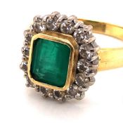 AN EMERALD AND DIAMOND CLUSTER RING. A CENTRAL TRAP CUT EMERALD IN A RUB OVER SETTING SURROUNDED