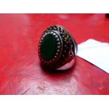 A GENTS OVAL CUT GREEN GEMSTONE SIGNET RING IN A CARVED SILVER BLACK AND GILDED HEAVY MOUNT.