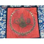 A BLACK CLOTH BACKED AND FRAMED RED SILK BANNER EMBROIDERED IN TWO GOLD COLOURED THREADS WITH A