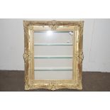GLAZED WALL DISPLAY CABINET FORMED AS AN ORNATE GILT PICTURE FRAME WITH GLASS SHELVES WITHIN,