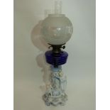 Oil lamp with blue glass reservoir below a white glass floral shade, supported by a porcelain column