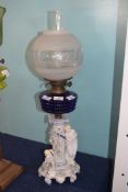 Oil lamp with blue glass reservoir below a white glass floral shade, supported by a porcelain column