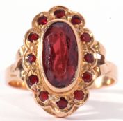 Mid-grade yellow metal dress ring, the oval panel centring a red stone in rub over setting