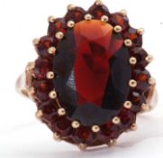 Garnet cluster dress ring, the large oval faceted garnet raised above small garnet surround, all