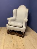 A large grey upholstered wing arm chair, with a Jacobean style wooden cross stretcher below, to