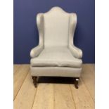 A large grey upholstered, high backed winged arm chair, raised on wooden legs