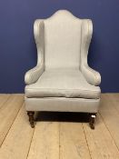 A large grey upholstered, high backed winged arm chair, raised on wooden legs