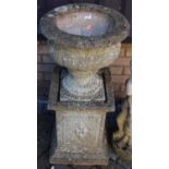 A reconstituted stone pedestal garden urn, set within a square high plinth (break to urn base),