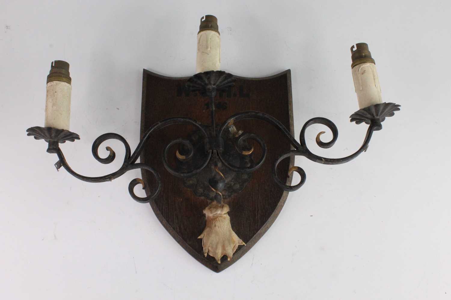 A mid-20th century wrought iron three sconce wall light fitting on an oak shield shaped plinth,