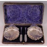 An Edwardian cased pair of silver shell butter dishes with accompanying knives, each dish of scallop