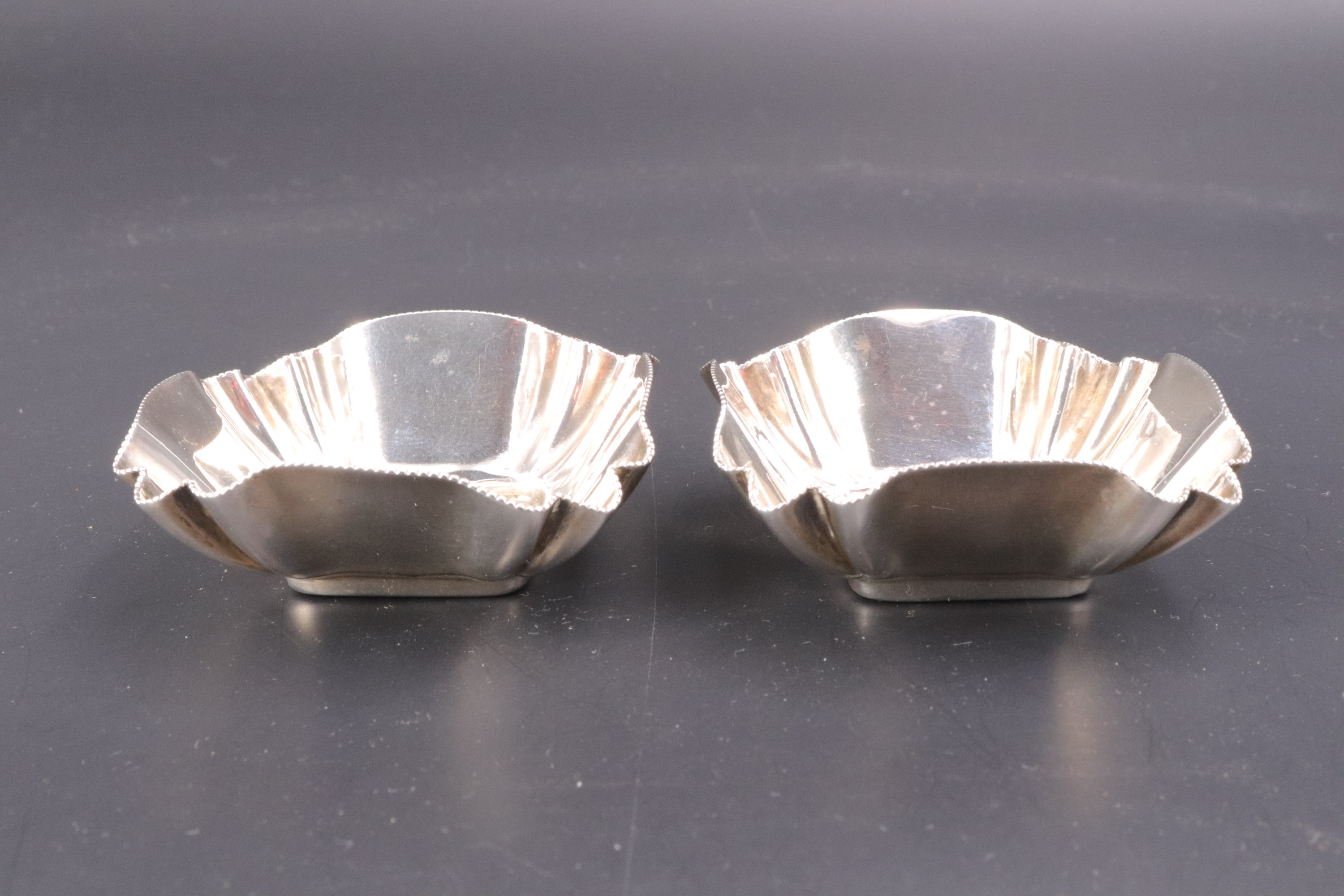 A pair of late Victorian silver salt cellars, each of cusped square section with a millegrain rim,