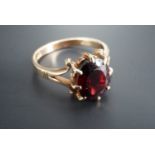 A 1970s garnet finger ring, the faceted oval stone of 9 mm x 7 mm claw-set between the scroll