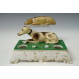 An early 19th century porcelain encrier, modelled with a recumbent greyhound behind two vacant
