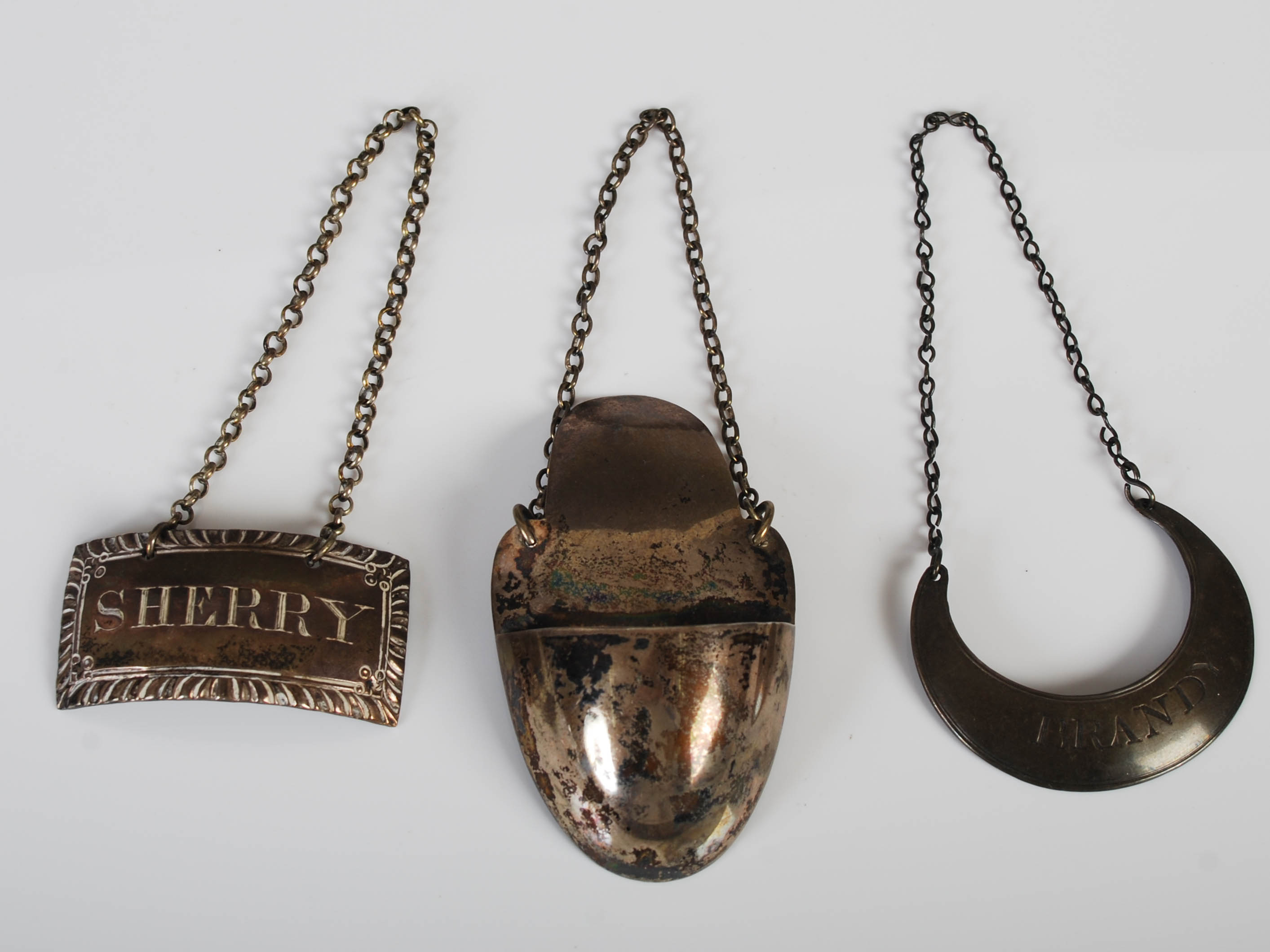 Two silver spirits labels and a silver drip catcher, the crescent shaped BRANDY label marked for