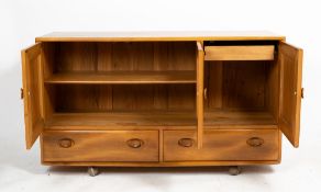 AN ERCOL LIGHT ELM SIDEBOARD with three panelled doors above two drawers, on rollable casters, 130cm