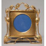 A GILT BRASS PHOTOGRAPH FRAME, C1910, OF SQUARE SHAPE WITH ROUND APERTURE BORDERED BY FLOWERS, ROD