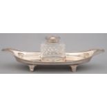 A GEORGE V SILVER MOUNTED OVAL CUT GLASS INKWELL ON CANOE SHAPED STAND, REEDED RIM AND FEET, 26CM L,