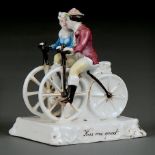 "KISS ME QUICK". A GERMAN PORCELAIN FAIRING, C1862-75, THE LOVERS EACH ASTRIDE A BONESHAKER BICYCLE,