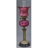 A Victorian oxidised brass columnar oil lamp, on black glazed earthenware foot, with cranberry glass