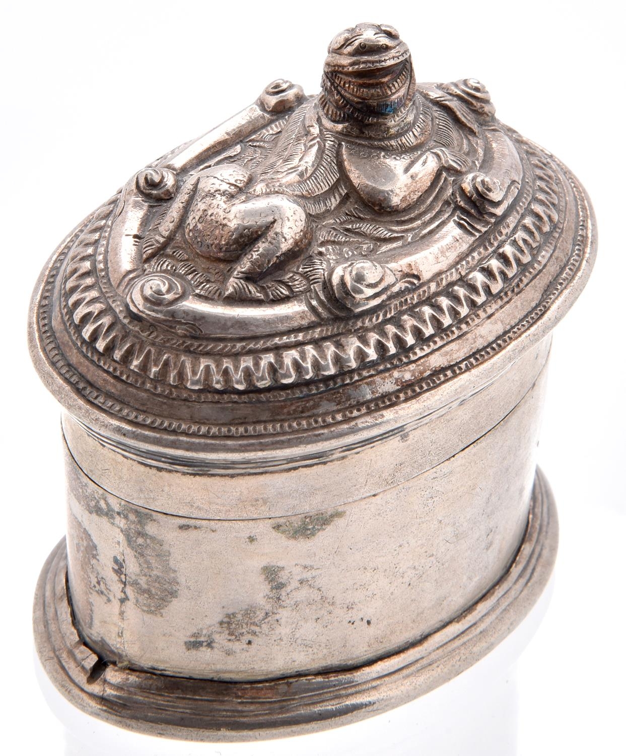 A Burmese oval silver lime box and repousse cover, early 20th c, the cover worked with a recumbent