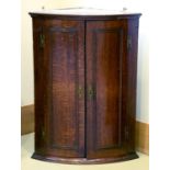 A George III bow fronted oak hanging corner cupboard, the pair of moulded doors with prominent