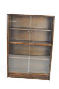 A modern, brown stained, glazed bookcase with three shelves behind sliding glass doors,