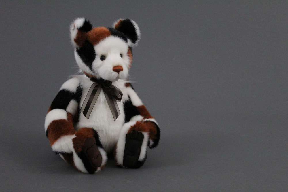 A soft plush Charlie Bear, named "Riddles", having patchwork patterned fur in tan, black and white,