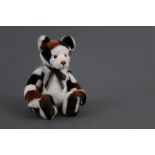 A soft plush Charlie Bear, named "Riddles", having patchwork patterned fur in tan, black and white,