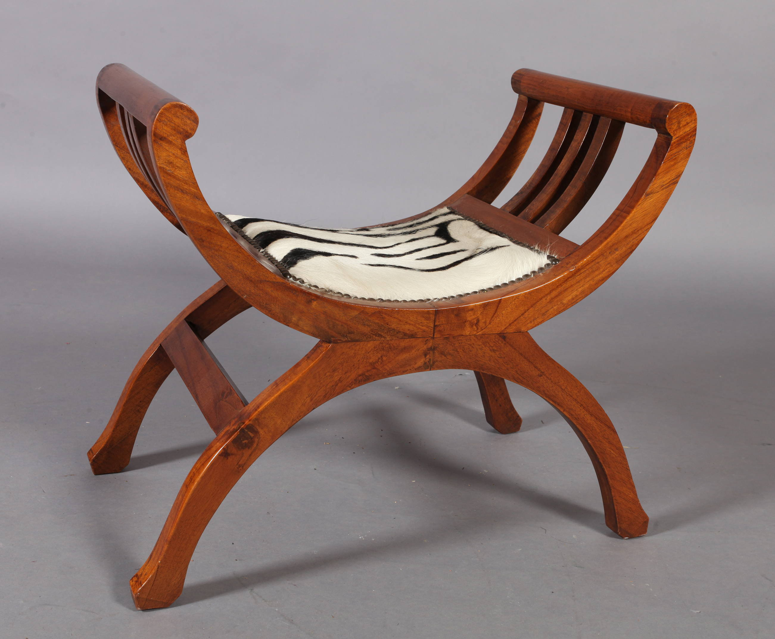 A reproduction hardwood x-frame stool, the seat upholstered with zebra skin within close brass