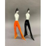 A pair of Cmielow china figures of girls in flared trousers, white and black and orange and black,