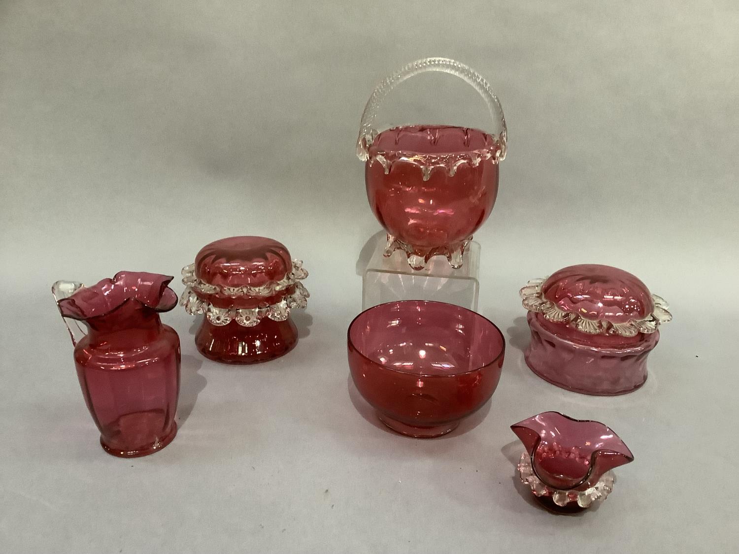 Six items of cranberry glass including two lidded jars with prunt rims together with a jug with