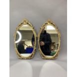 A pair of gilt metal framed oval wall mirrors each with a basket of flowers cresting, tassle and