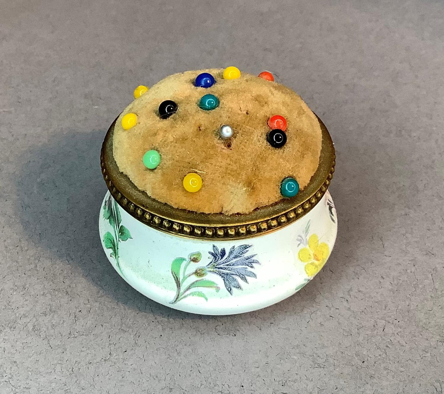 A brass mounted circular enamel pin cushion, ogee form body printed flowers beneath a beaded brass