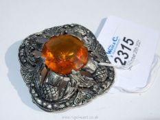 A white metal brooch set with a 24 mm diameter faceted and polished amber coloured stone surrounded