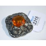 A white metal brooch set with a 24 mm diameter faceted and polished amber coloured stone surrounded