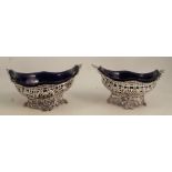 A pair of pierced silver bonbon dishes, with scroll edge, blue glass liners, raised on scroll bases,