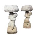 A pair of composition stone urns with leaf moulded bowls supported by seated cherubs on naturalistic