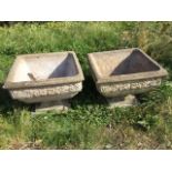 A pair of square composition stone urns with moulded rims above floral embossed friezes, raised on